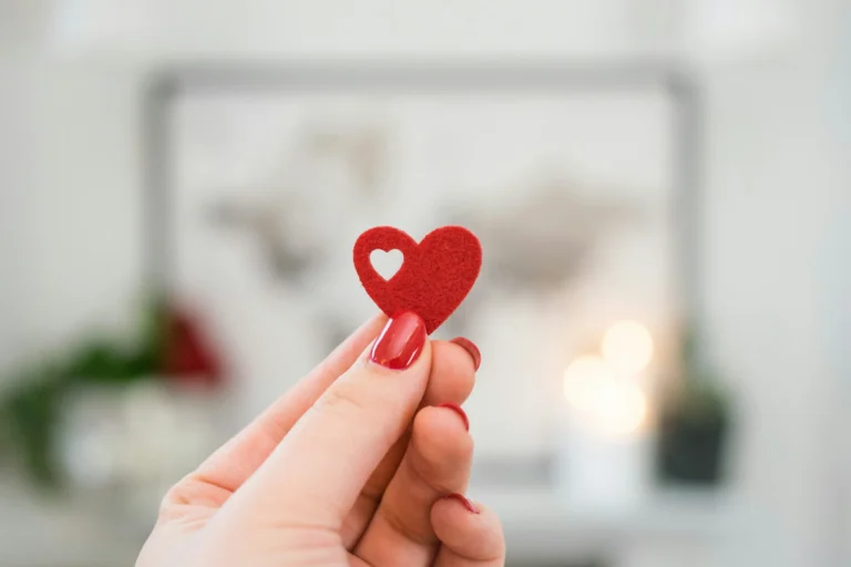 In the picture there is the hand of a woman with red polished nails holding small red heart made of felt. The felt has also a cut shaped heart inside of it.