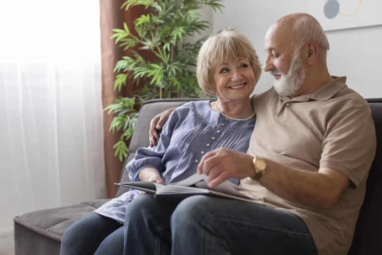 In the picture there is an old couple sat on a sofa browsing through a magazine. The woman is watching the man's face and smiles. On the back there is a green plant.