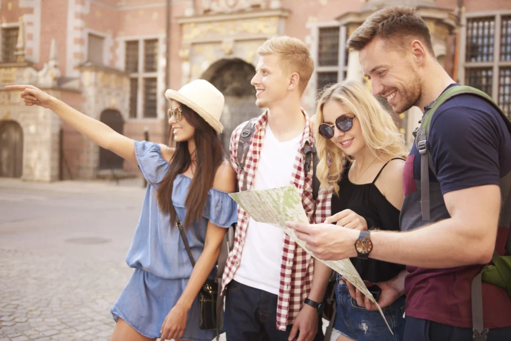 The picture shows a group of people, two men and two women, with summer clothes visiting a city. A girl indicates a far away point to one of the guy, while the other two people are checking out the map.